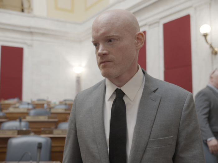 A man in a gray suit and black tie stands in the West Virginia House of Delegates chamber. Behind him, rows of empty hardwood desks with placards and microphones sit.