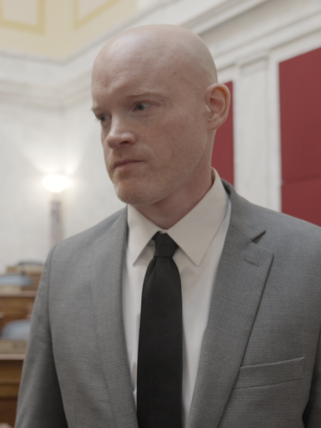 A man in a gray suit and black tie stands in the West Virginia House of Delegates chamber. Behind him, rows of empty hardwood desks with placards and microphones sit.