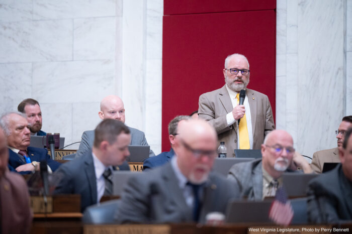 Rows of men sit at hardwood desks behind laptops and microphones. A man in a brown jacket and yellow tie is standing up at one desk, speaking into a handheld microphone and looking forward.