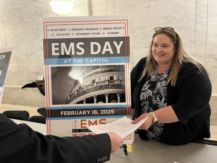 A woman stands behind a table displaying a sign that reads "EMS Day at the Capitol." She is handing a packet of paper to someone on the other side of the table, whose face is out of view.