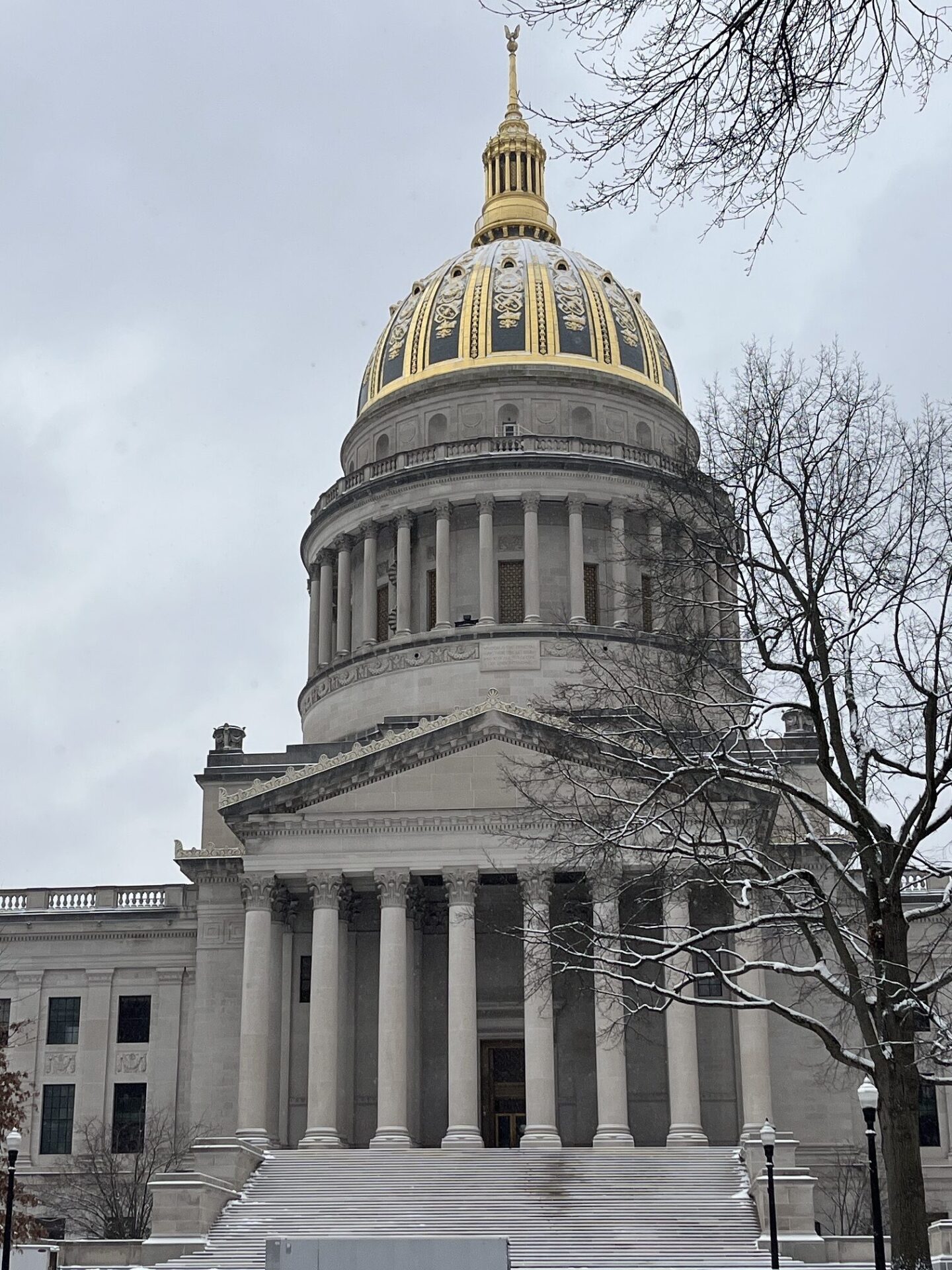 The golden dome of the State Capitol is covered in a light layer of snow. Snow lines a sidewalk down below, leading to stairs and the tall columns and windows of the State Capitol building.
