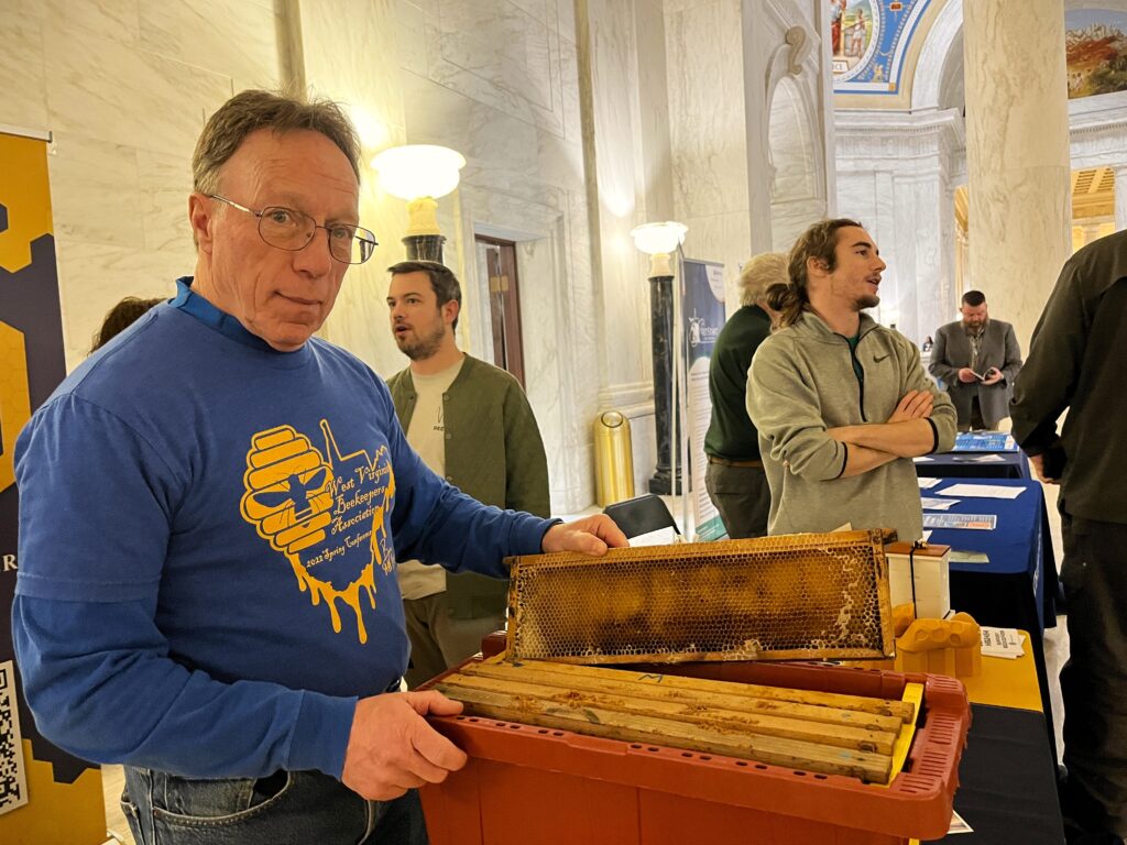 A man with glasses and a blue shirt pulls racks of honeycomb out of a red tub. He is looking into the camera. The tub is placed atop a table in a row of tables, and people stand behind him on other sides of the tables, talking and observing materials atop them.