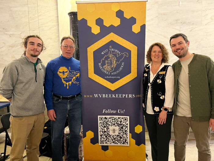 Two people stand on either side of an upright sign that depicts honeycombs and the outline of West Virginia. Text on the sign reads "West Virginia Beekeepers Association." Behind the sign is a white marble wall of West Virginia's State Capitol.