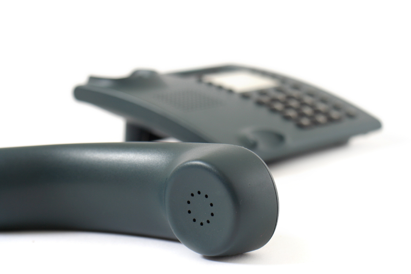 The receiver of a black office phone sits in the foreground in front of the body of the phone on a white setting and white backdrop.