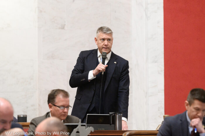 A man in a suit speaks into a microphone. Beside him sit several other people in formal attire seated at hardwood desks. Behind him is a white marble wall.