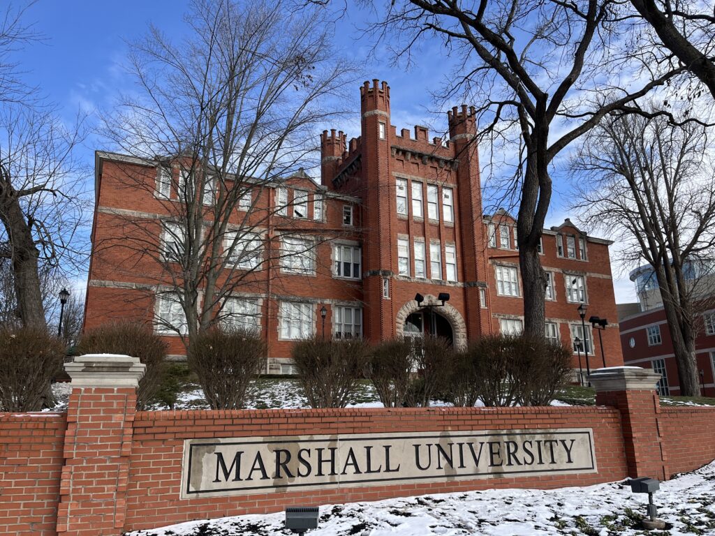 A sign reads "Marshall University" in the garden of a red brick building with a tall stone archway, several windows and four spires atop the center area of the building.