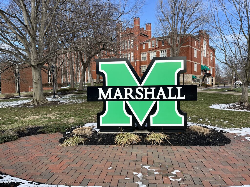 A large, upright sculpture depicts a green letter "M" with the letters "Marshall" in white text written across it. The sculpture is at the center of a garden surrounded by a brick walkway, and old brick buildings are visible behind it.