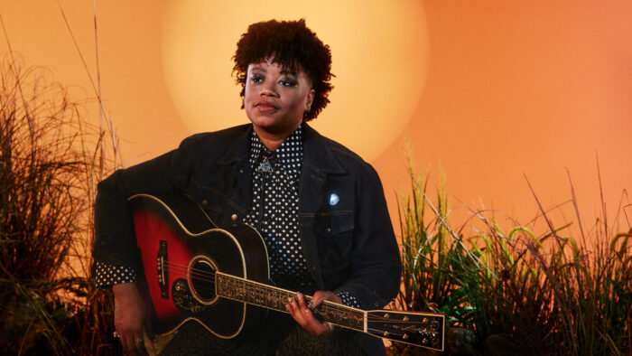 A person holding a guitar and looking to their left into the distance. They are sitting in front of an orange backdrop surrounded by plants.