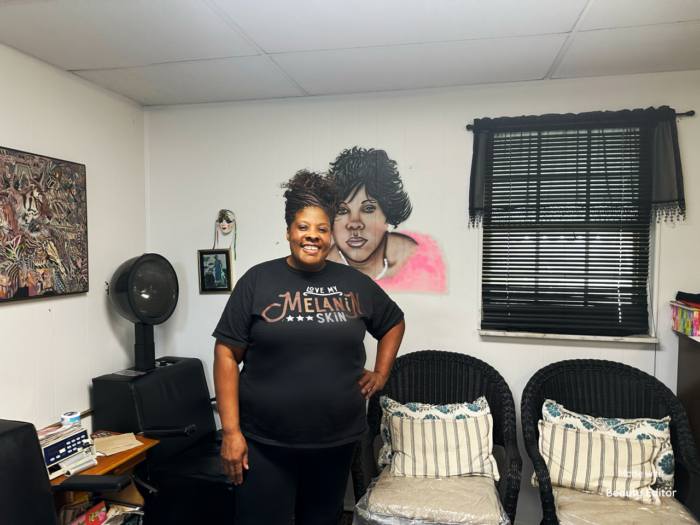 A woman standing in a room in front of mural of a woman painted on the wall. A black salon chair with a hair dryer is on her left and two black chairs on her right.