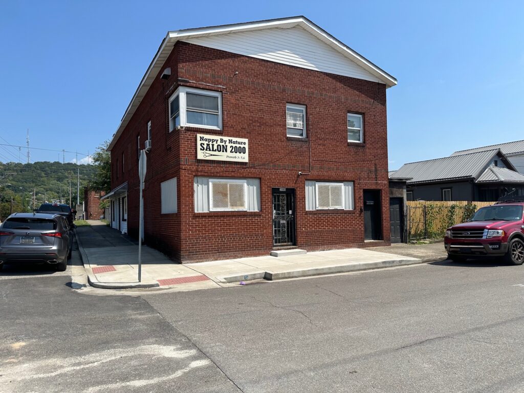 A red brick building with a sign that says "Nappy By Nature SALON 2000" and has a large image of scissors. The building is on a street and the sky is blue.