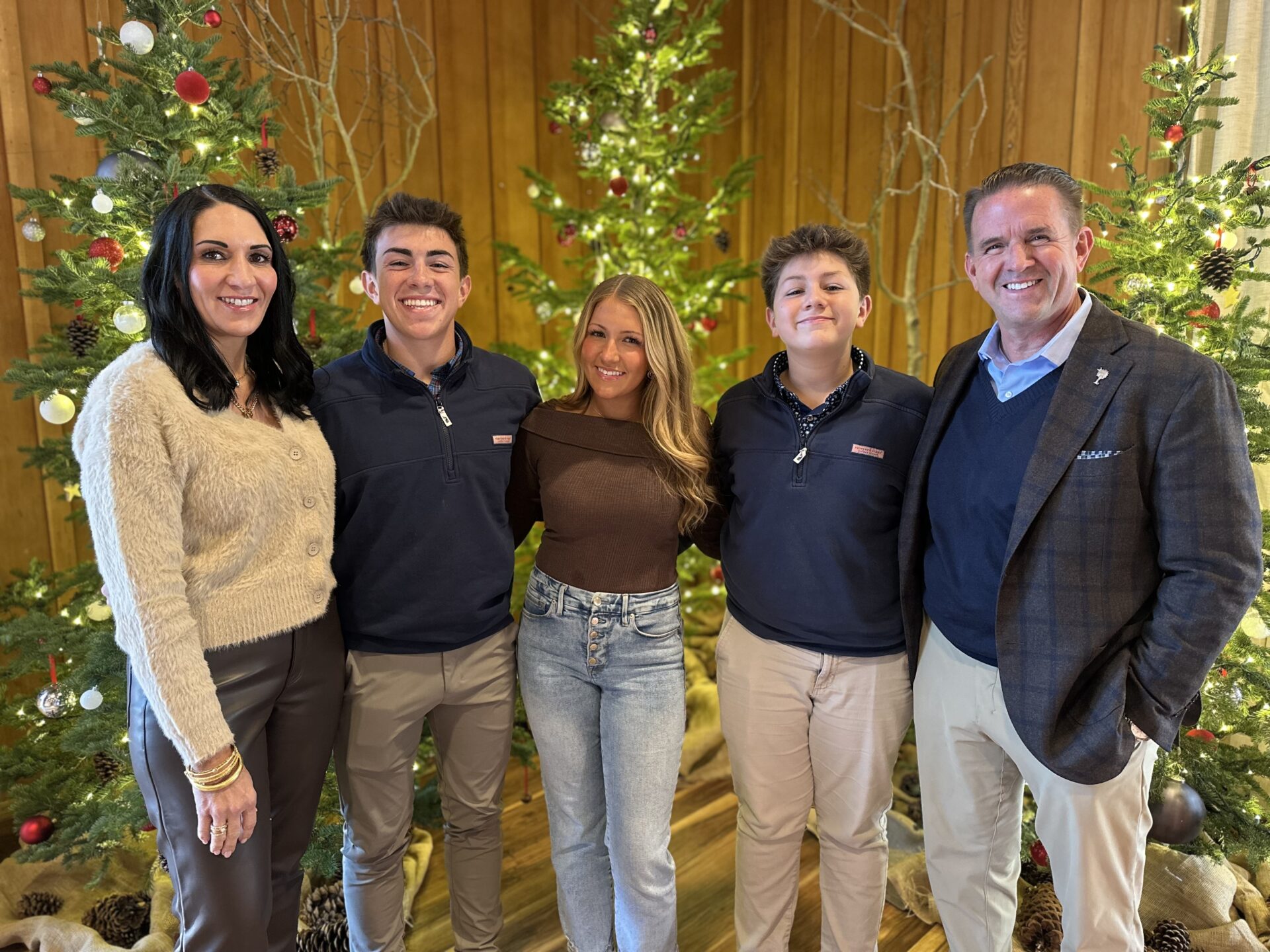 A family of five stands in a row in front of simple Christmas decorations in front of a wood paneled wall