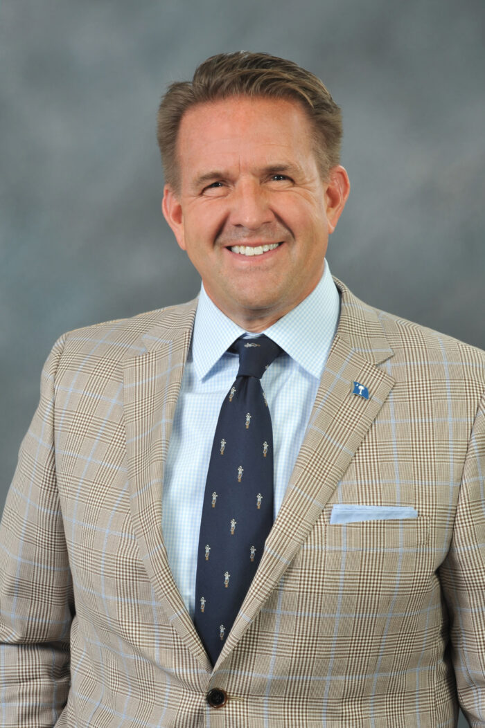 A man in a tan sports jacket with a light blue checker pattern worn over a white shirt and a dark blue tie.