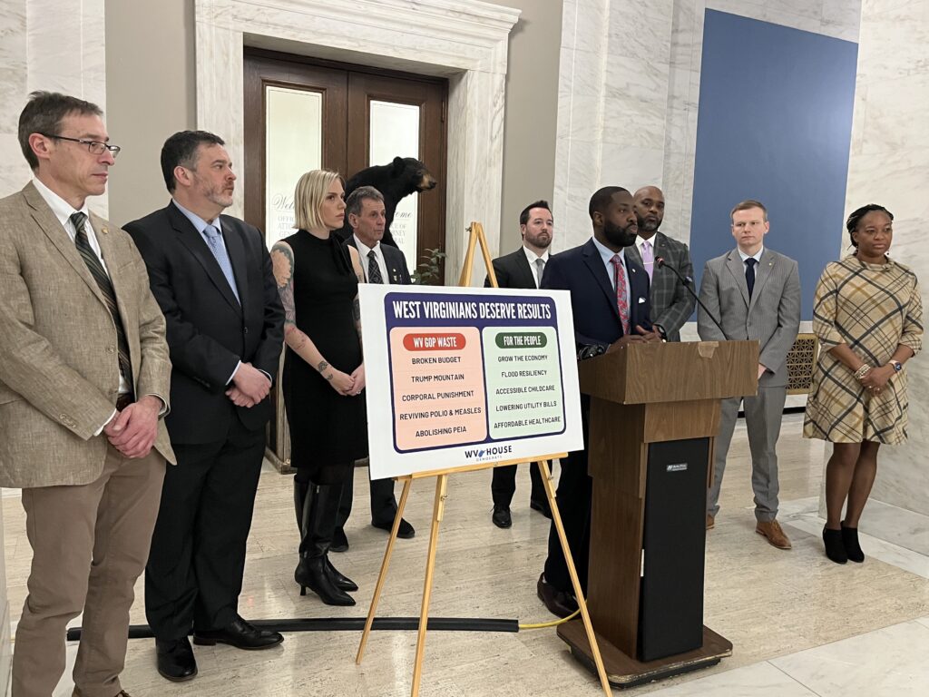 A group of people in formal attire stand around a podium. One man in a suit stands at the podium, speaking into a microphone. Beside him is a sign with two lists: one labeled "WV GOP Waste," and the other labeled "For The People."