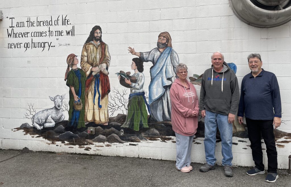 three people stand by a wall that depicts Jesus talking to children. It says on the wall I am the bread of life. Whoever comes to me will never go hungry..."