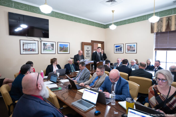 A wide shot of a conference room shows various people, almost all wearing suits, sitting at long wood tables arranged in a U-shape. Two men in the center of frame stand at lecterns near a door.