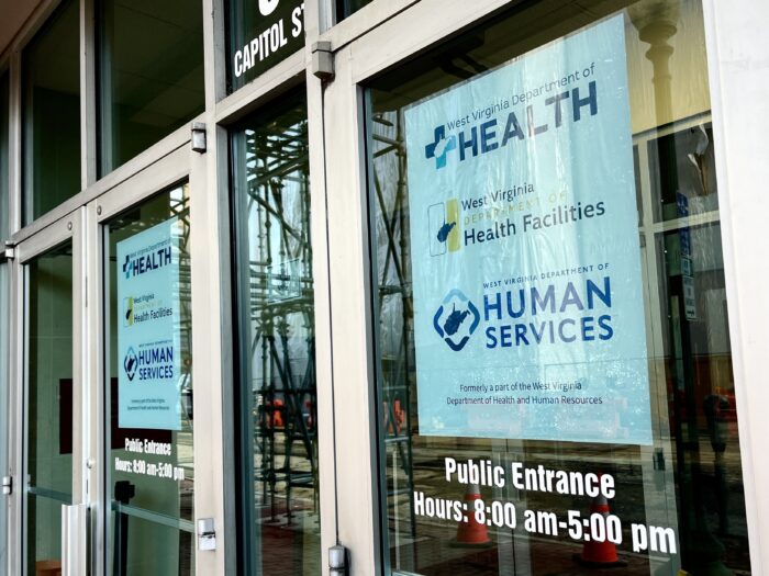 A glass-pane door displays two of the same sign on either side, showing the logos of the West Virginia Department of Health, West Virginia Department of Health Facilities and West Virginia Department of Human Services. Beneath the side, white text reads "Public Entrance, Hours: 8 a.m. to 5 p.m."