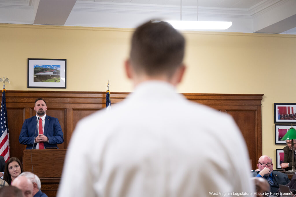 A man wearing a black suit and red tie speaks from a podium opposite the view of the photographer, who stands over the shoulder of a physician wearing his white coat.