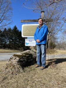 A man wearing a blue jacket and jeans stands next to a sign in a yard.