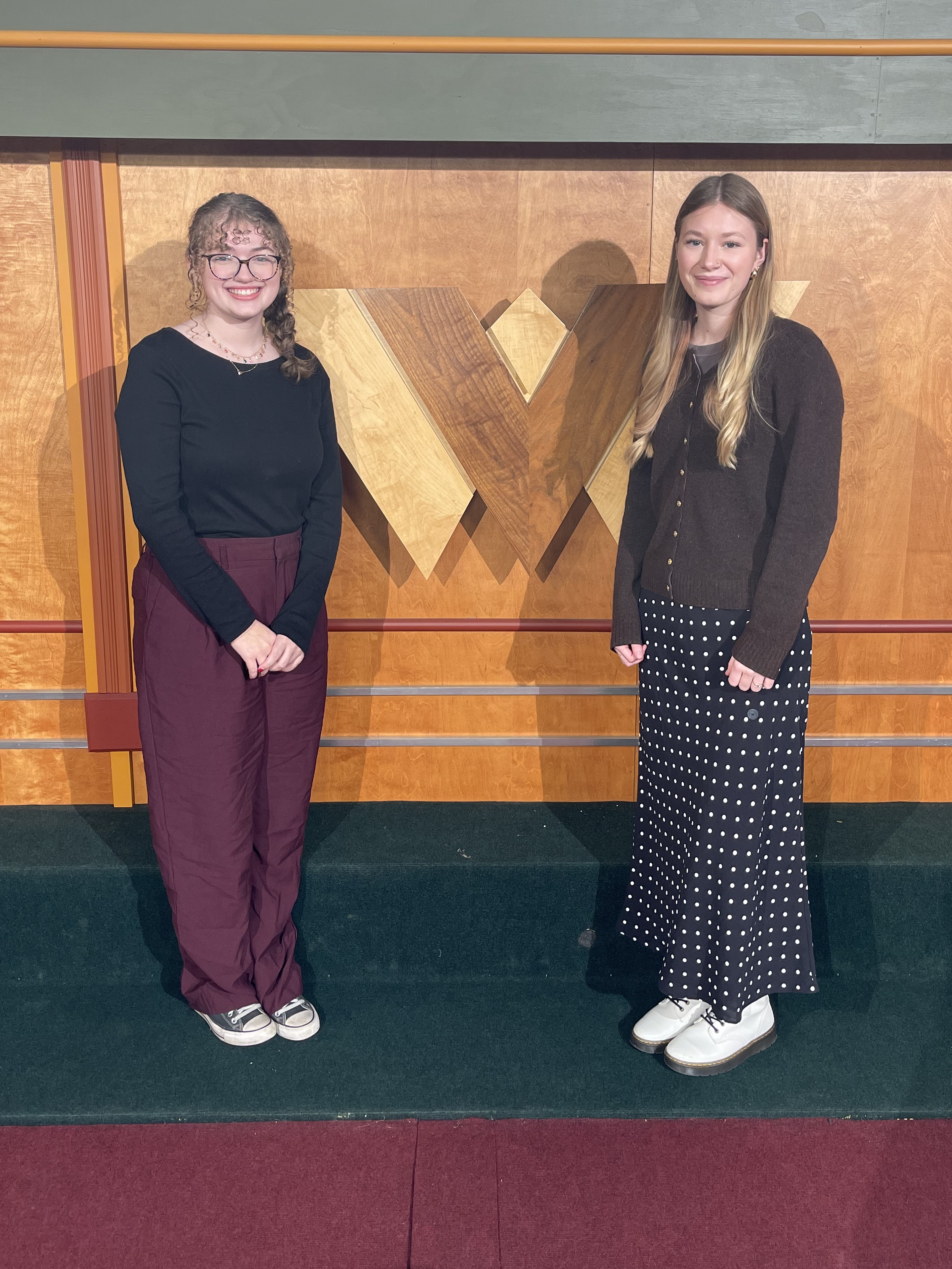 Two young women stand next to each other smiling. They are on a sound stage. Behind them is the WVPB logo.