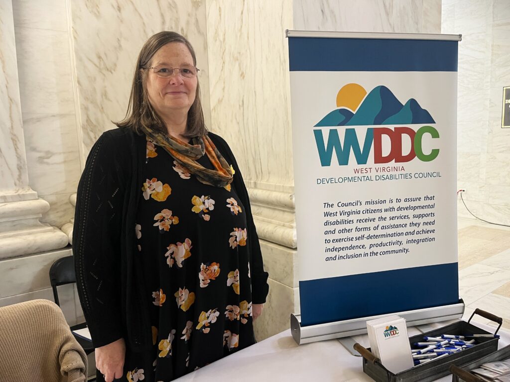 A woman in glasses, a black cardigan and a floral print dress stands behind a table, which displays an upright sign reading "West Virginia Developmental Disabilities Council." Surrounding the table, the walls and floor of the room are made from white marble.