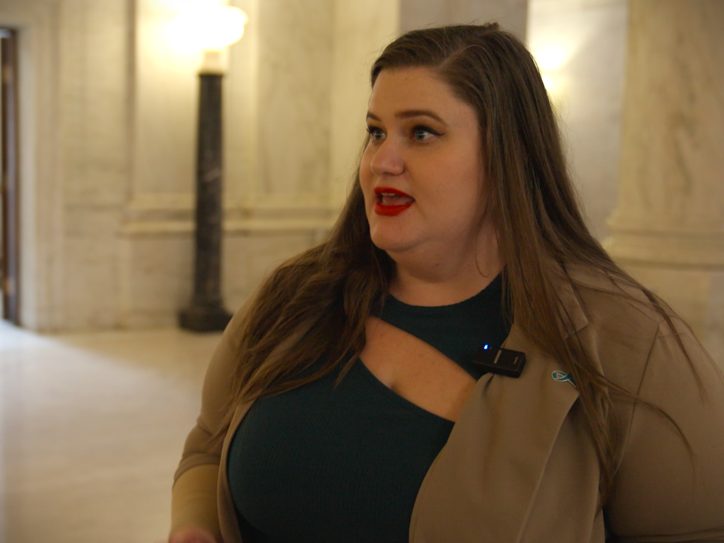 A woman in a blazer looks away from the camera, talking to someone off screen. She stands in a white marble hallway.