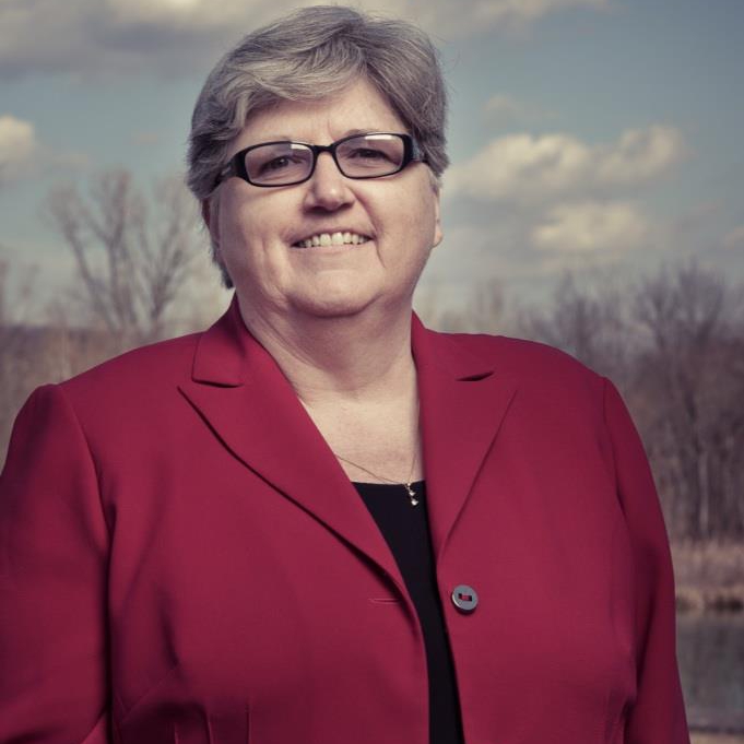 A woman with short cropped hair, black glasses and a red suit jacket looks toward the camera, smiling. Behind her, a backdrop of barren trees and a partially cloudy sky is visible.
