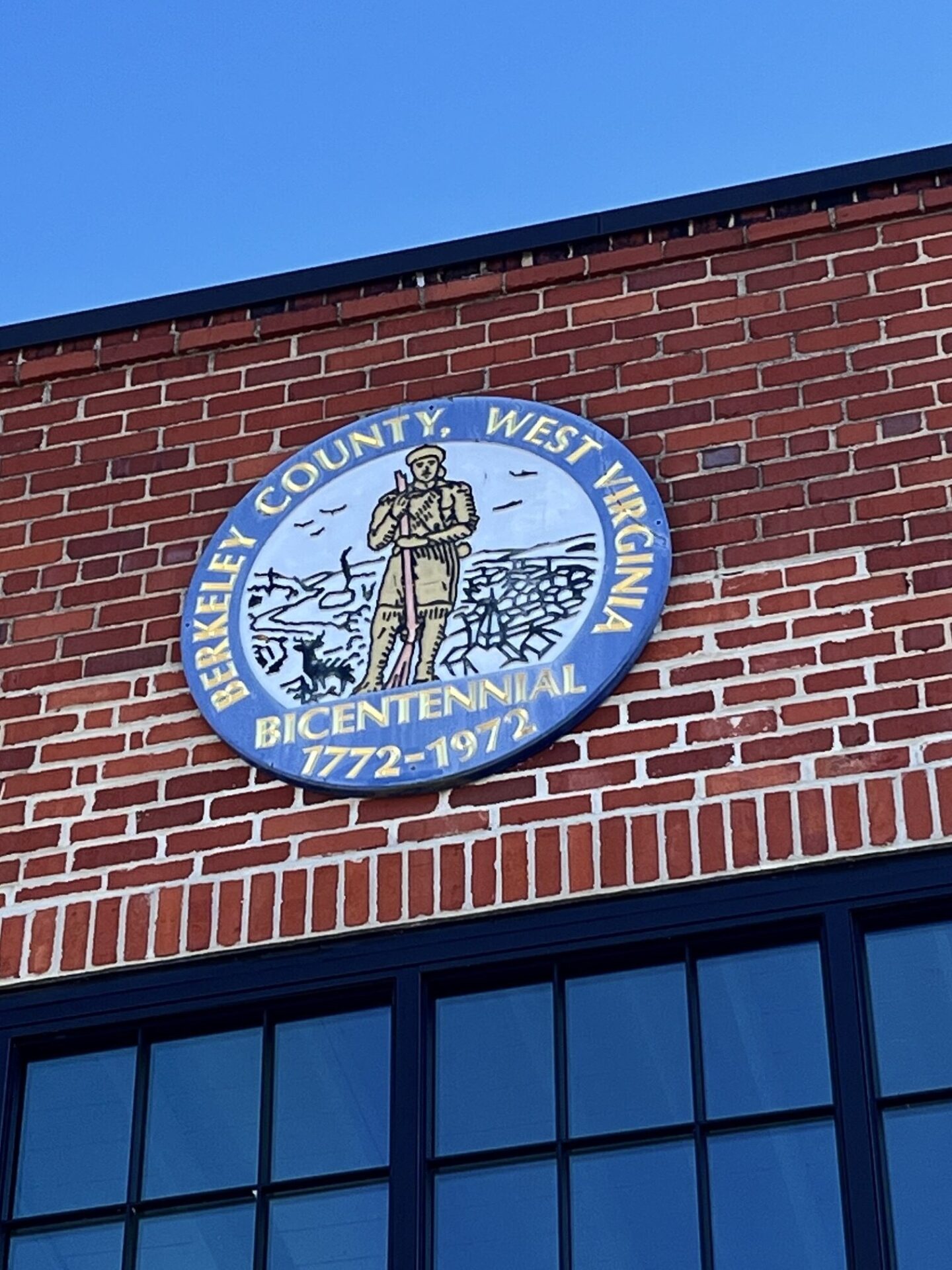 A blue and yellow metal, circular seal is embedded onto the top of a red brick building, directly above a window pane. It shows a hunter holding a rifle and reads "Berkeley County, West Virginia Bicentennial, 1772-1972."