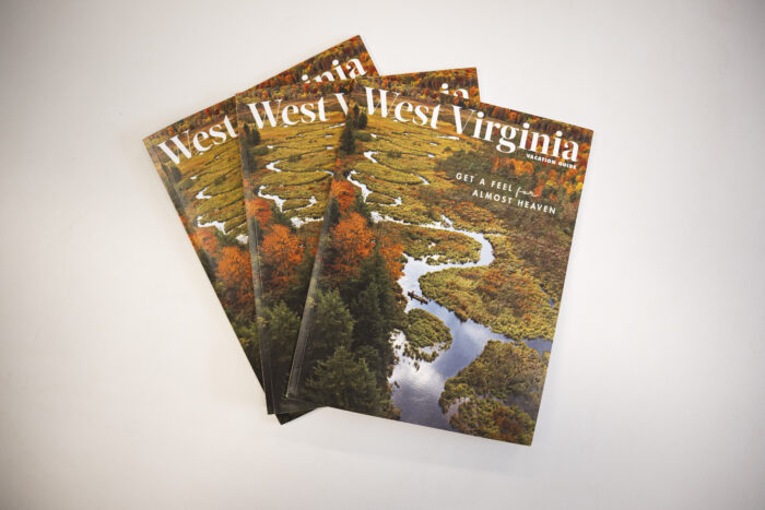 Picture shows a gray background. Three magazines with the words "West Virginia" and "Get A Feel for Almost Heaven" are displayed against a scenic aerial photo of fall trees with some orange leaves along a winding river.