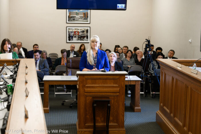 a woman with bright blond hair and a blue jacket stands at a pulpit with a audience behind her. She is facing the U-shaped desks around her, with her back to audience. 