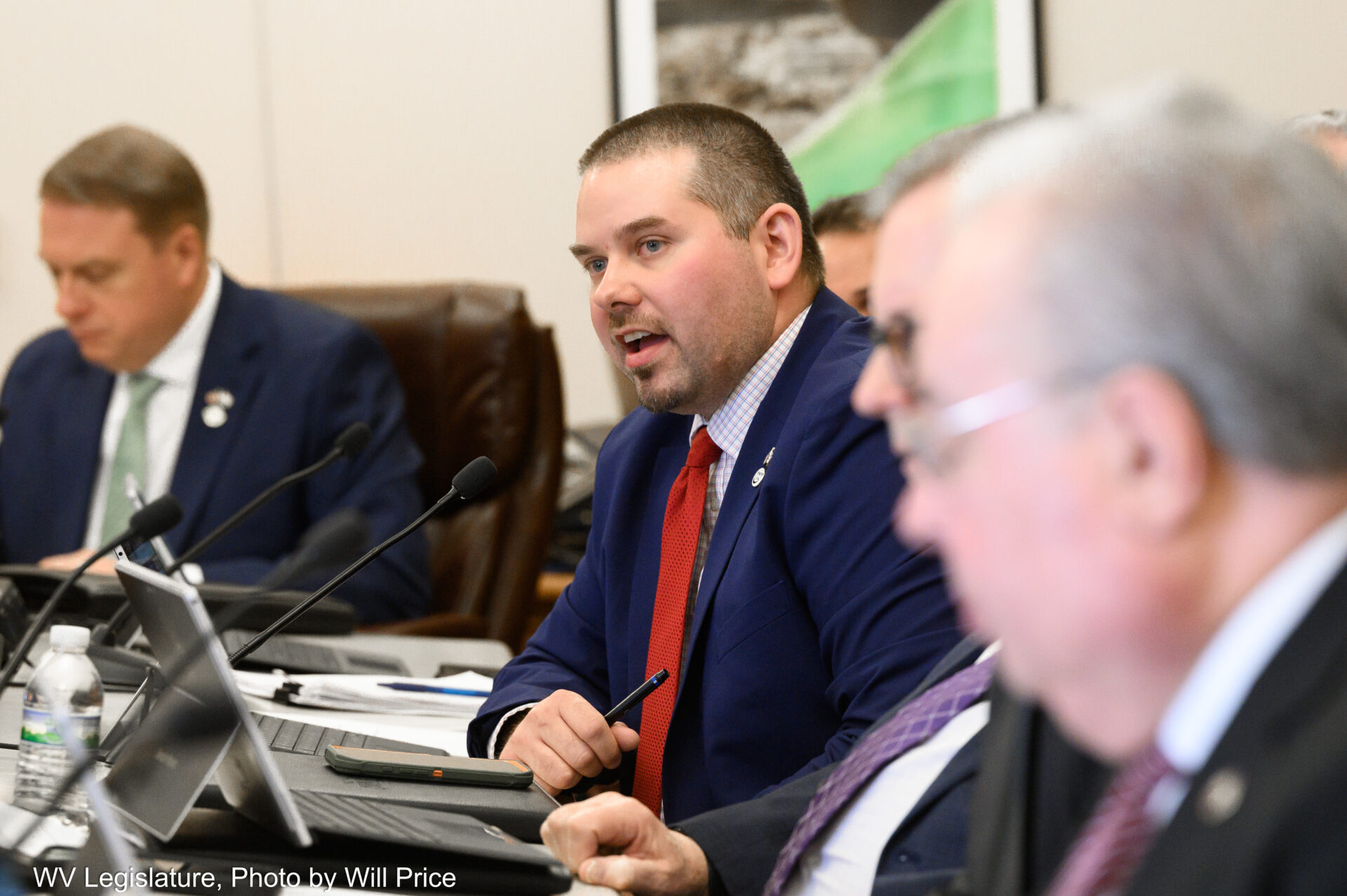 A man talks at desk, with a blurred man in the forefront of the photo.