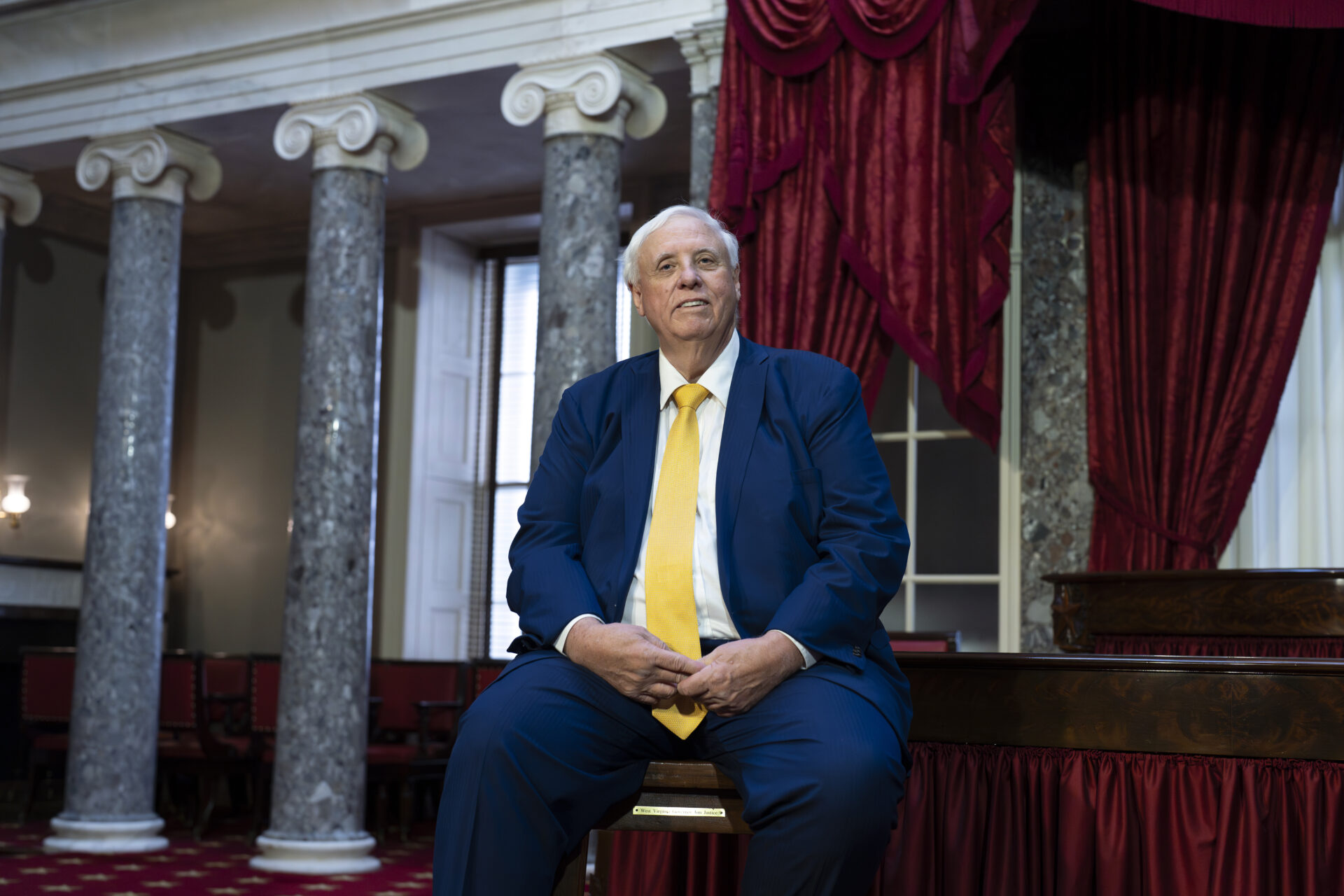 A large. white-haired man in a blue suit with a gold tie sits smiling on a desk in front of two tall, ornate columns and a window with red drapes.