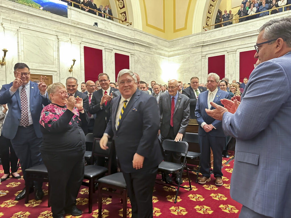 A man in a suit enters a crowded, ornate room as people in professional attire applaud.