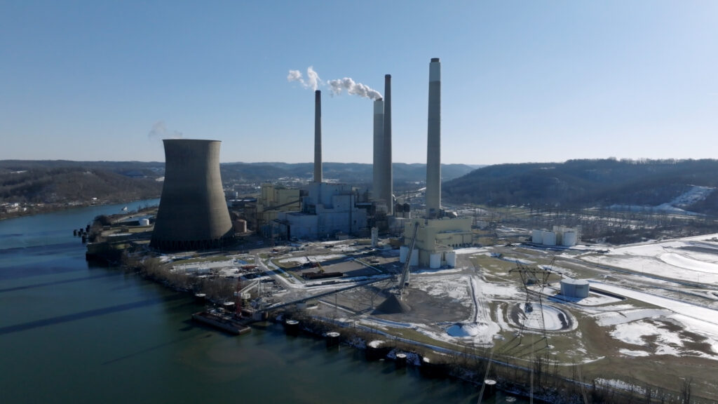 A large industrial facility with multiple stacks and concrete cooling towers looms over a snow-covered riverbank on a clear winter day.