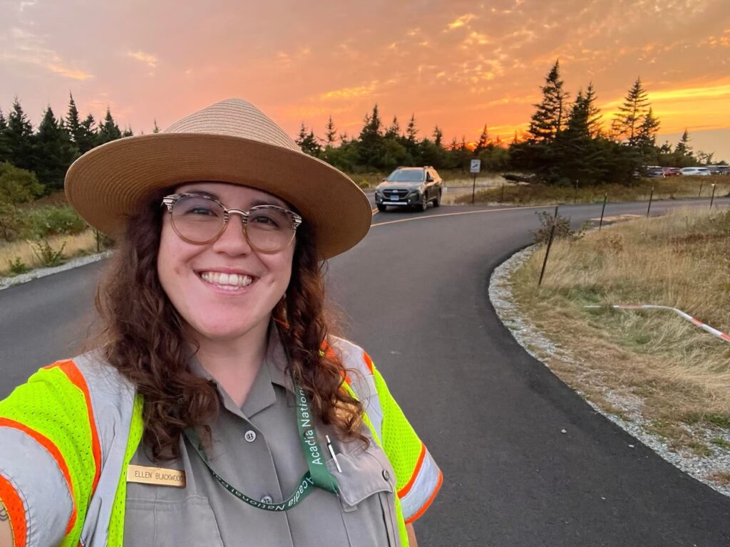 A young woman with dark, shoulder length hair wearing a park ranger uniform with a tan, broad-rimmed hat, stands along a highway with a sunset and tall pine trees in the background.