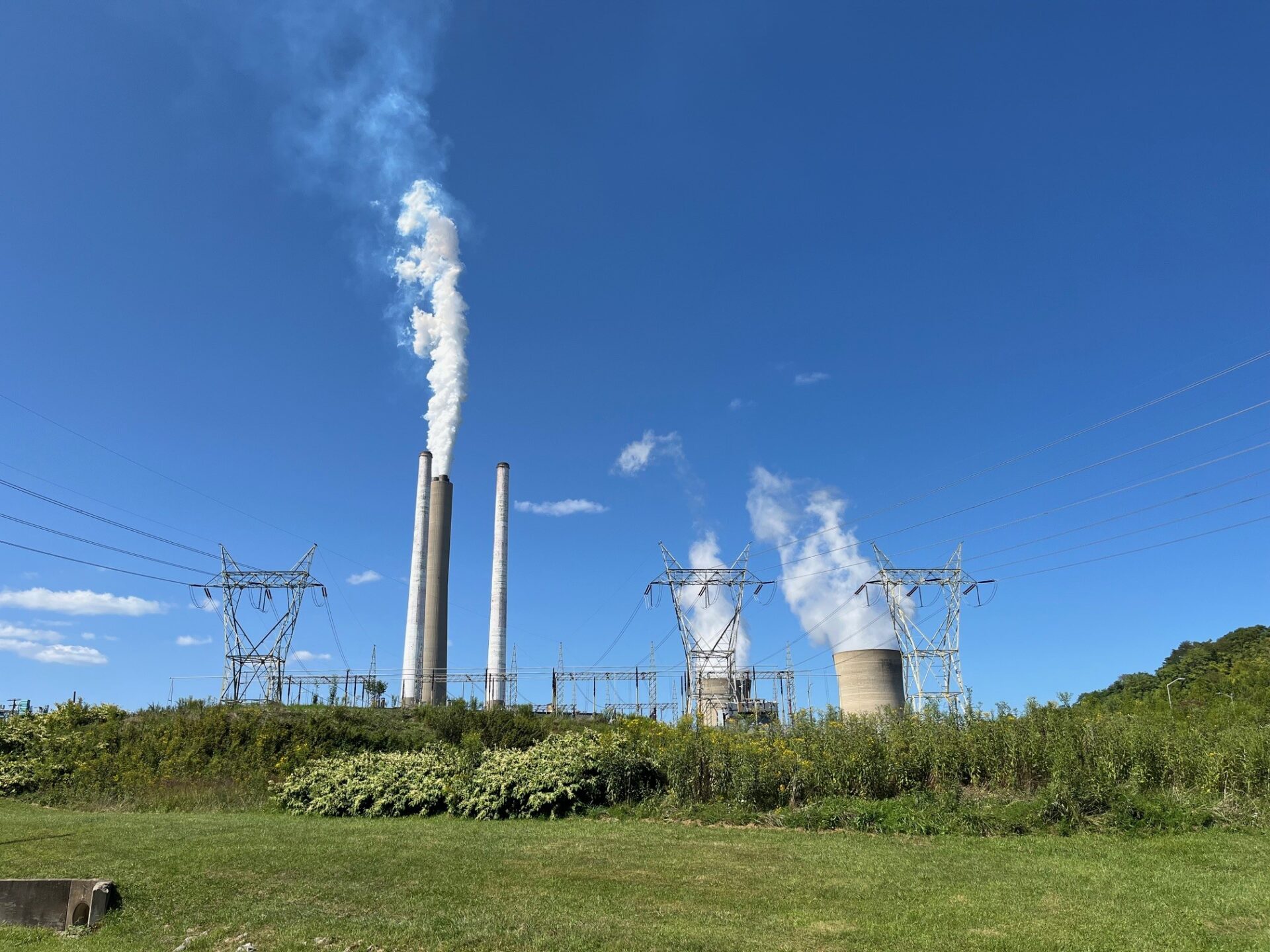 Puffs of steam rises from a stack and concrete cooling towers with a clear blue sky and green foreground.