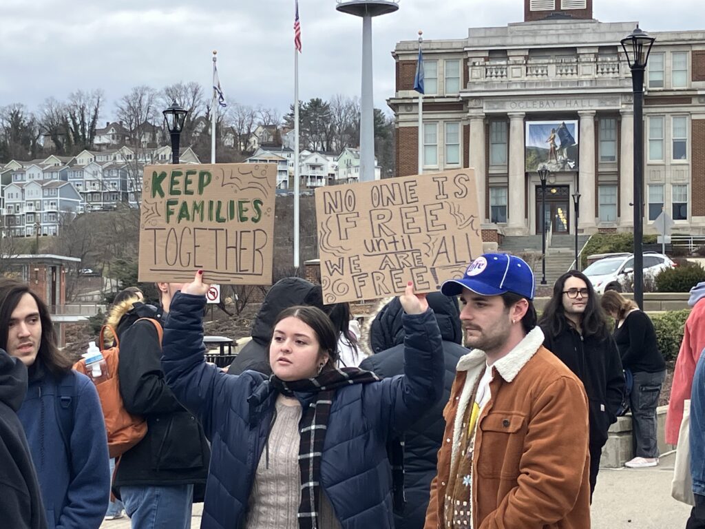 A young woman wearing a blue puffy jacket over a tan sweater holds up two signs. The sign on the left reads “Keep Families Together” and the sign on the right reads “No One Is Free Until We Are All Free” She stands below an academic building in the neoclassical style, with a columned facade below a grey sky