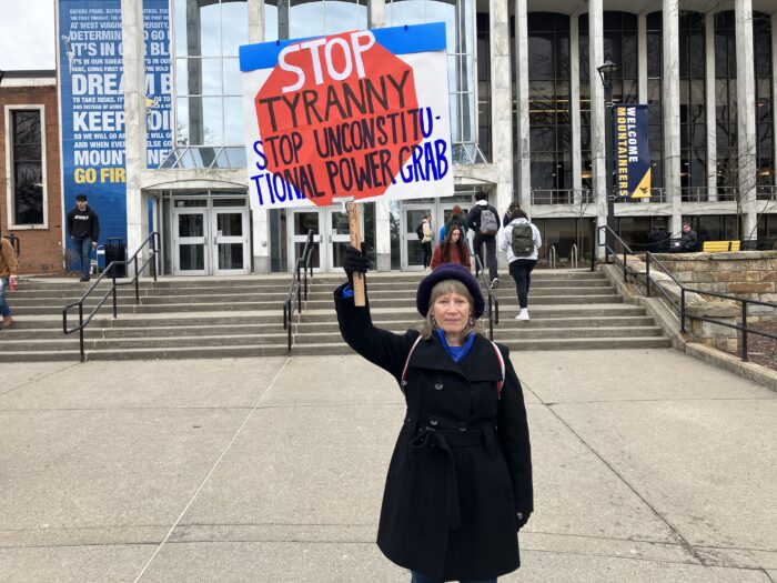 A woman wearing a fuzzy black bucket hat and a black jacket and black gloves over a royal blue shirt holds a large sign by a handle over her head. The sign reads “Stop Tyranny Stop Unconstitutional Power Grab” over a red octagon on a white field.