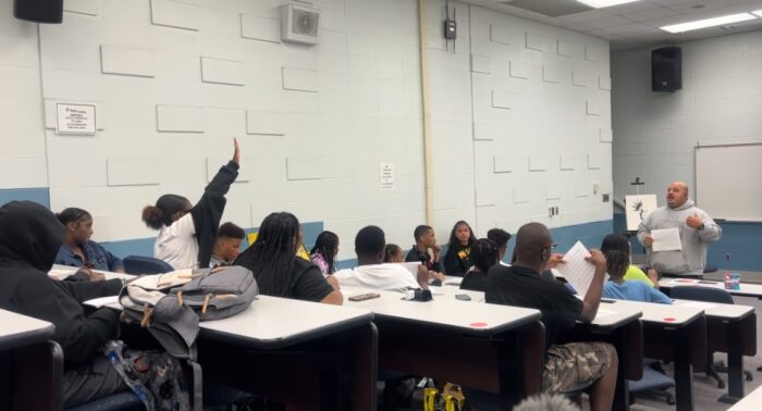 A girl raises her hand from near the back of a classroom full of students.