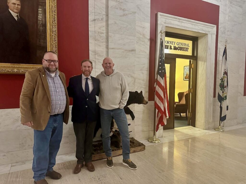 Three men stand togethr in a marble hallway against an ornate red backdrop with a large portrait to the left, an American flad and the entry to the W.Va. Attorney General's office to the right.