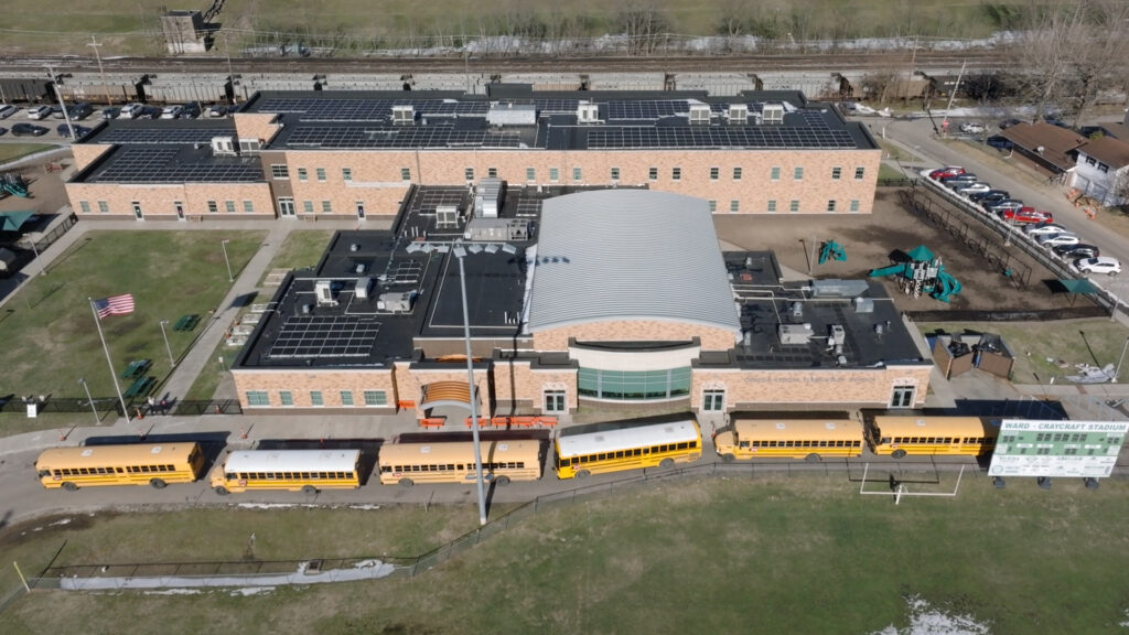 The roof of a school building, as viewed from a drone, with hundreds of solar panels on it, with a line of yellow school buses in front and empty railcars behind.