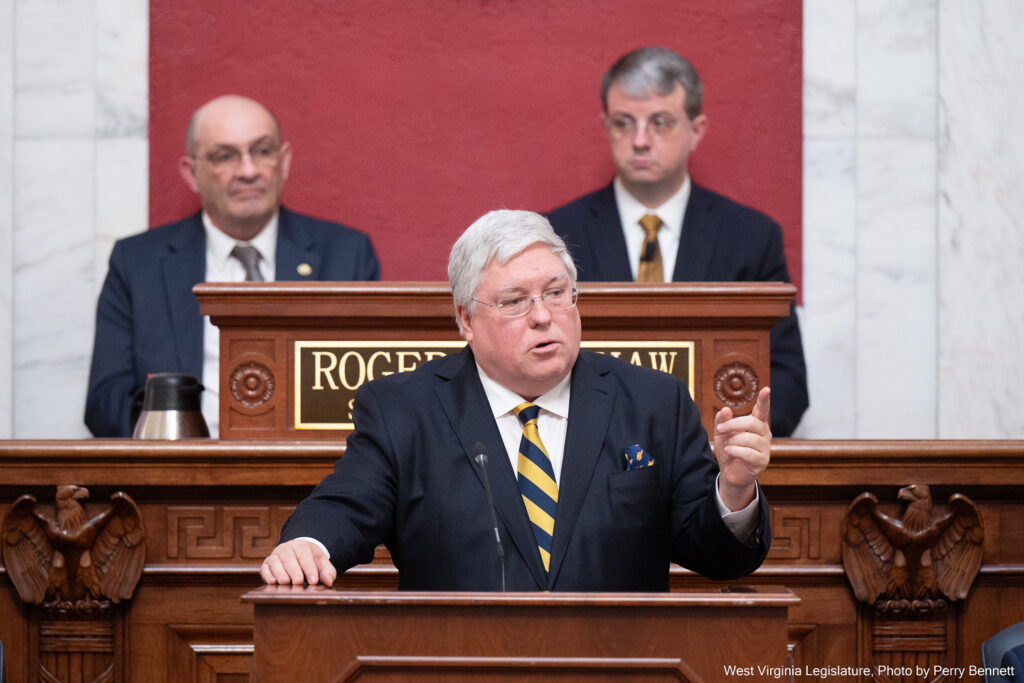 A white-haired man in a dark suit with a navy and gold striped tie speaks at a podium as two seated men in professional attire look on from behind him.