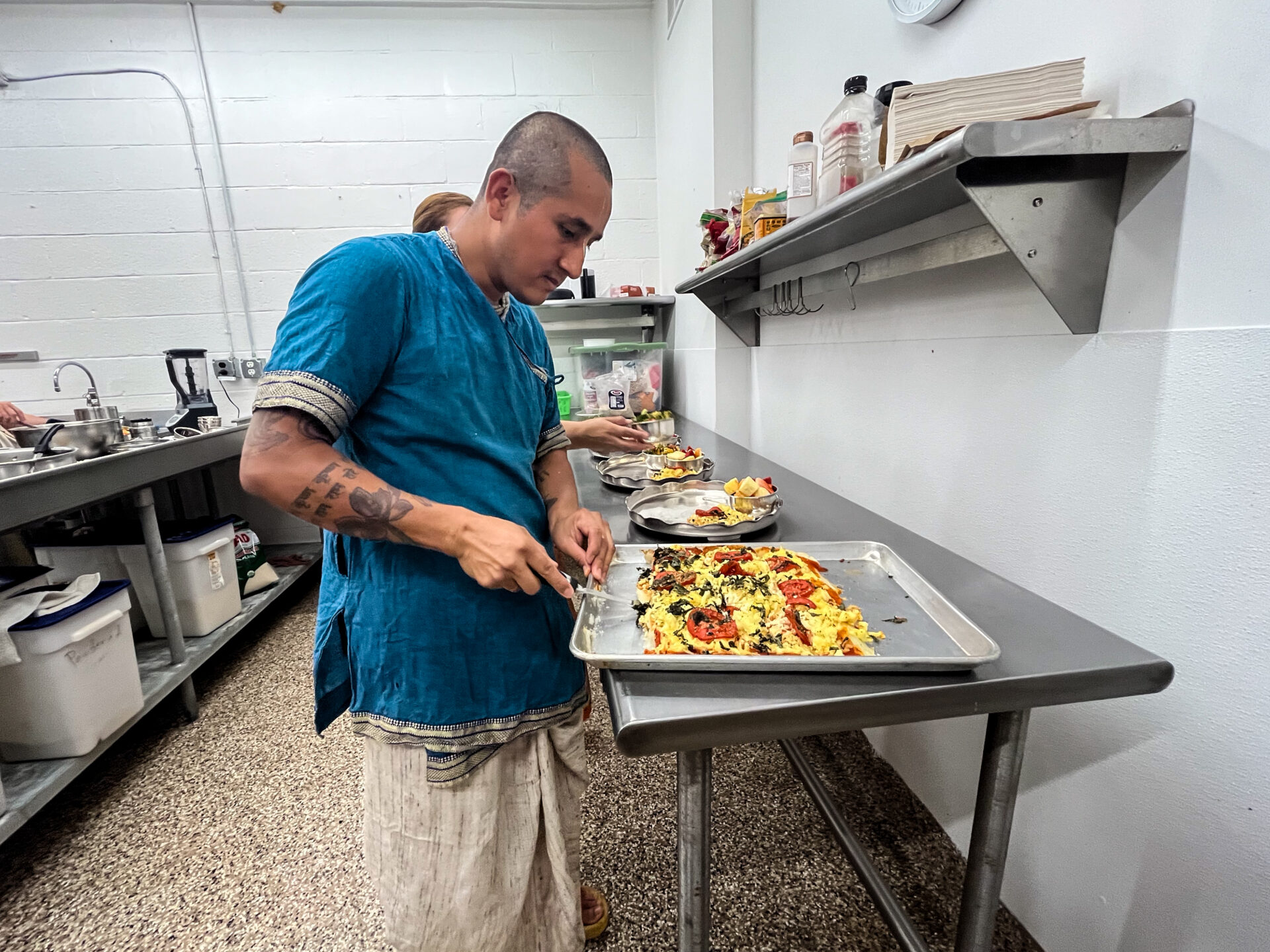 A man prepares food with red tomatoes, greens and other ingredients on top of a metal counter.