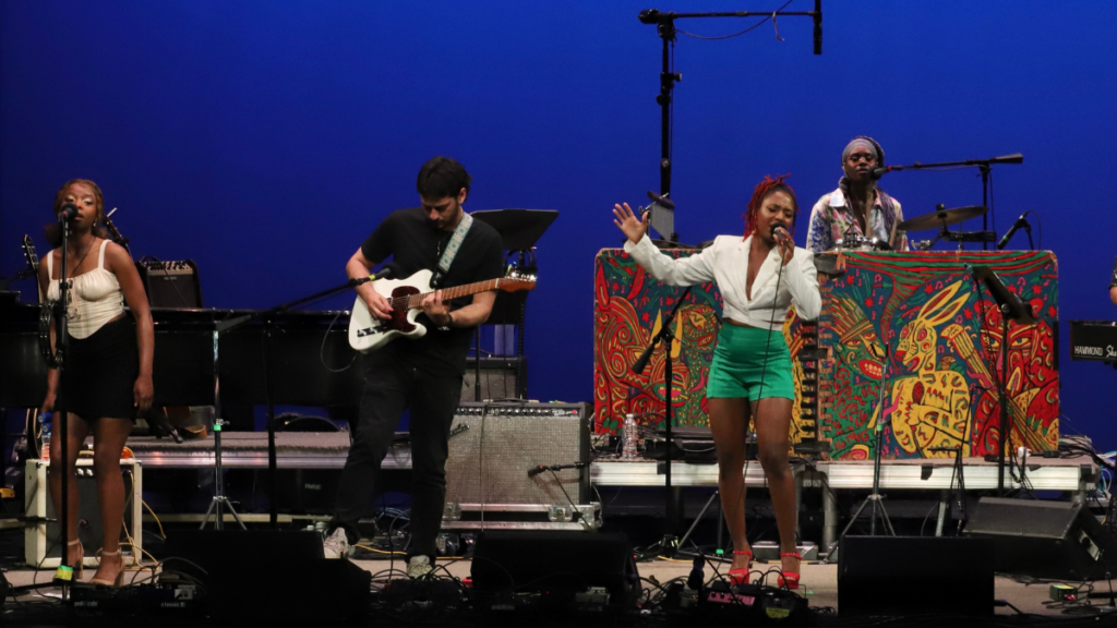 Four musicians, a woman on guitar, a man on guitar, a woman on drums, and a woman singing into a mic. The woman singing wears a white shirt and teal pants. She sings into a microphone.