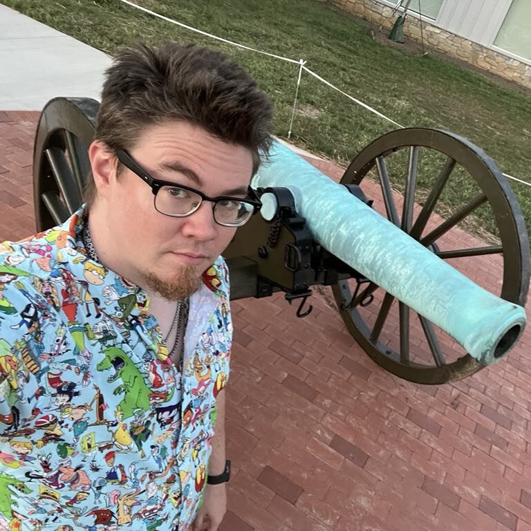 A man in a collared polo shirt with cartoon characters printed on it stands beside a historic-looking canon and looks into the camera for a selfie.