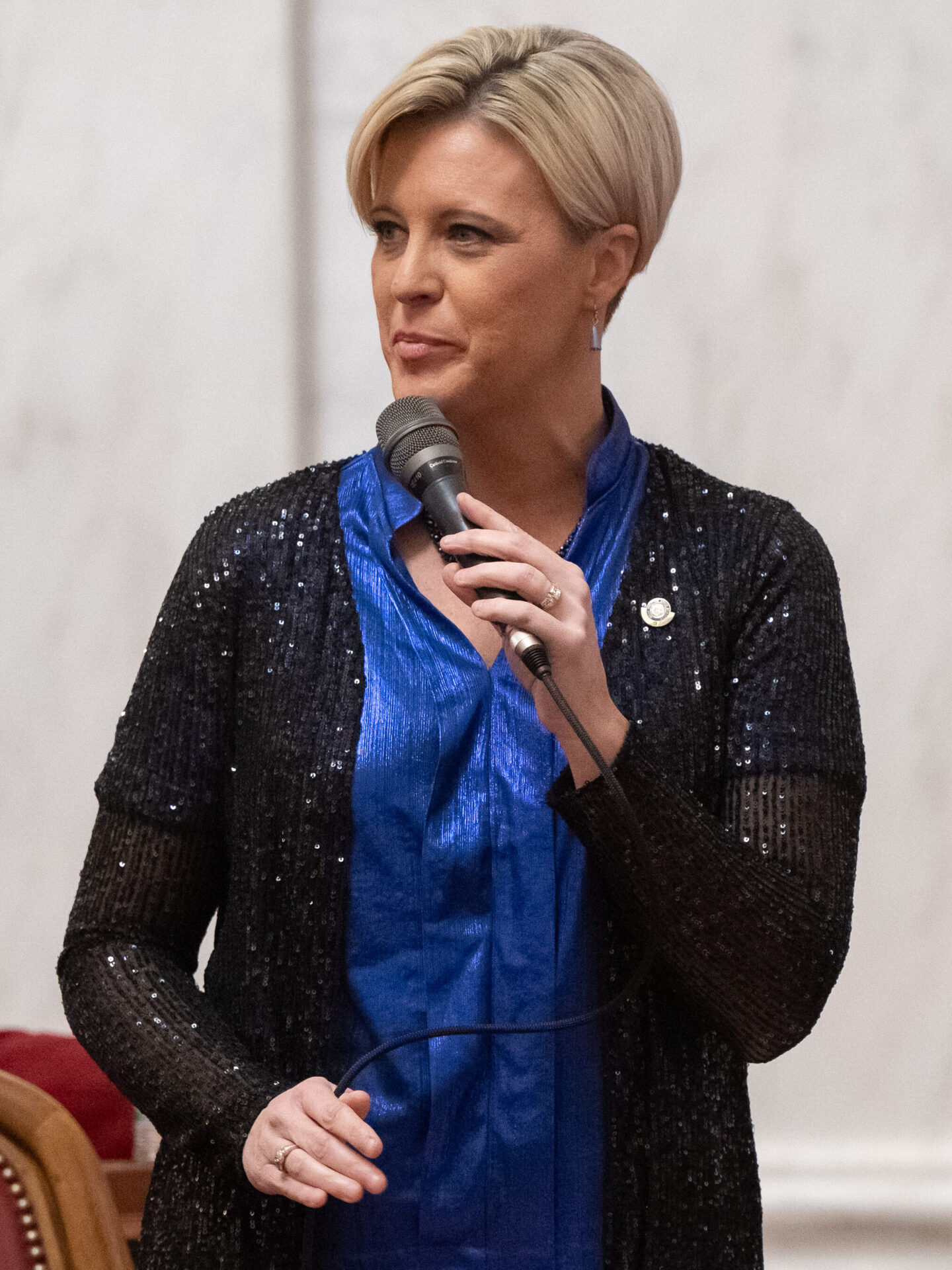 A woman weaking a sparkling knee length sheer black sweater over a satin royal blue blouse holds a microphone while standing in front of a chair with red leather lining. Behind her can be seen a woman seated at a wooden desk wearing a burgundy blazer over a white blouse. In the background a white marble wall has a column facade.