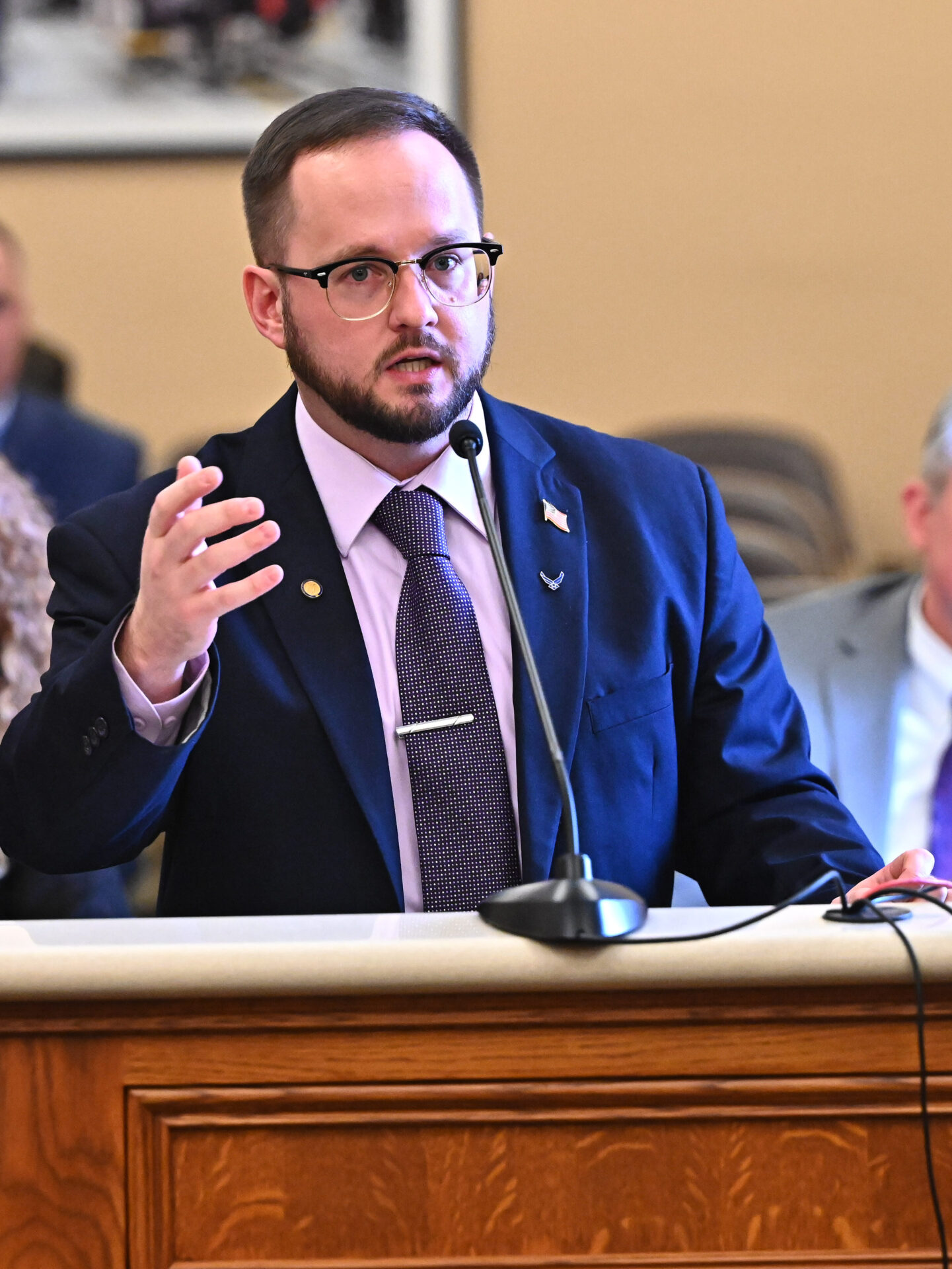 A man wearing glasses and a purple tie over a pink shirt with a navy blue suit gestures with his right hand while speaking at a lectern. On either side of him can be seen people seated at a table, to the right a man in a white shirt and purple tie with grey blazer and to the left a woman wearing glasses and a black blouse. Above the speaker's head, in the background, can be seen a framed photograph.