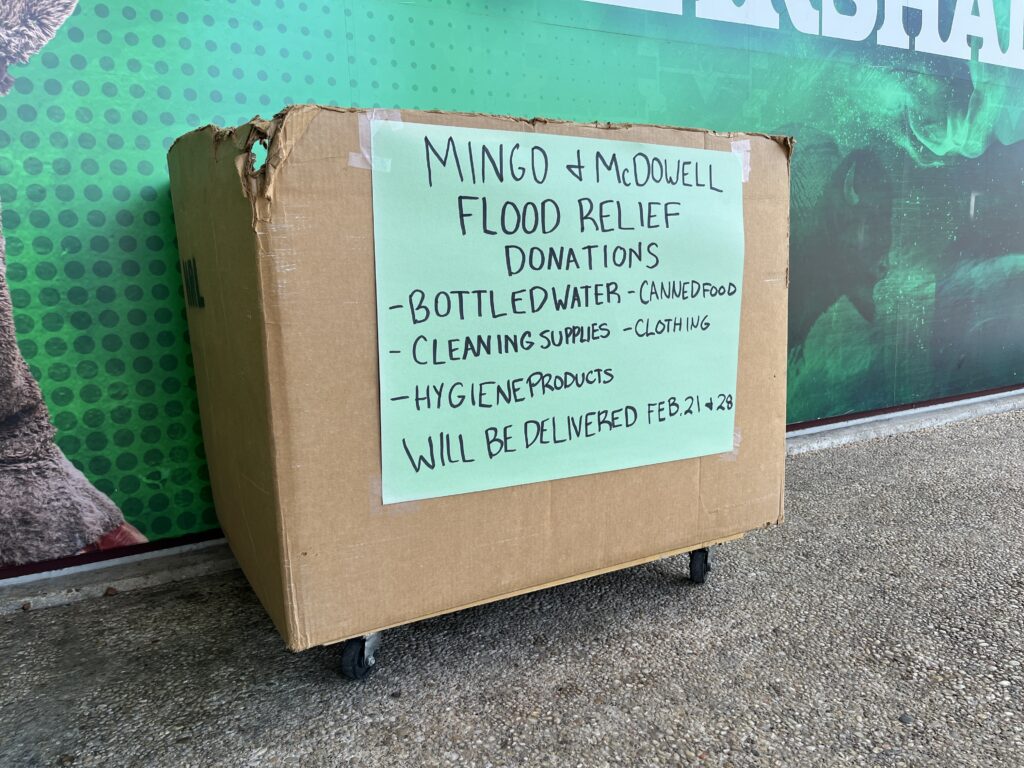 A cardboard box placed outside a building has a green handwritten sign taped on it, which reads: "Mingo and McDowell Flood Relief Donations."