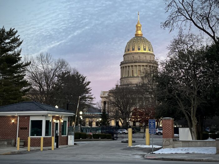 The sun sets behind the dome of the West Virginia State Capitol, viewed from a street in front of its parking lot entrance.