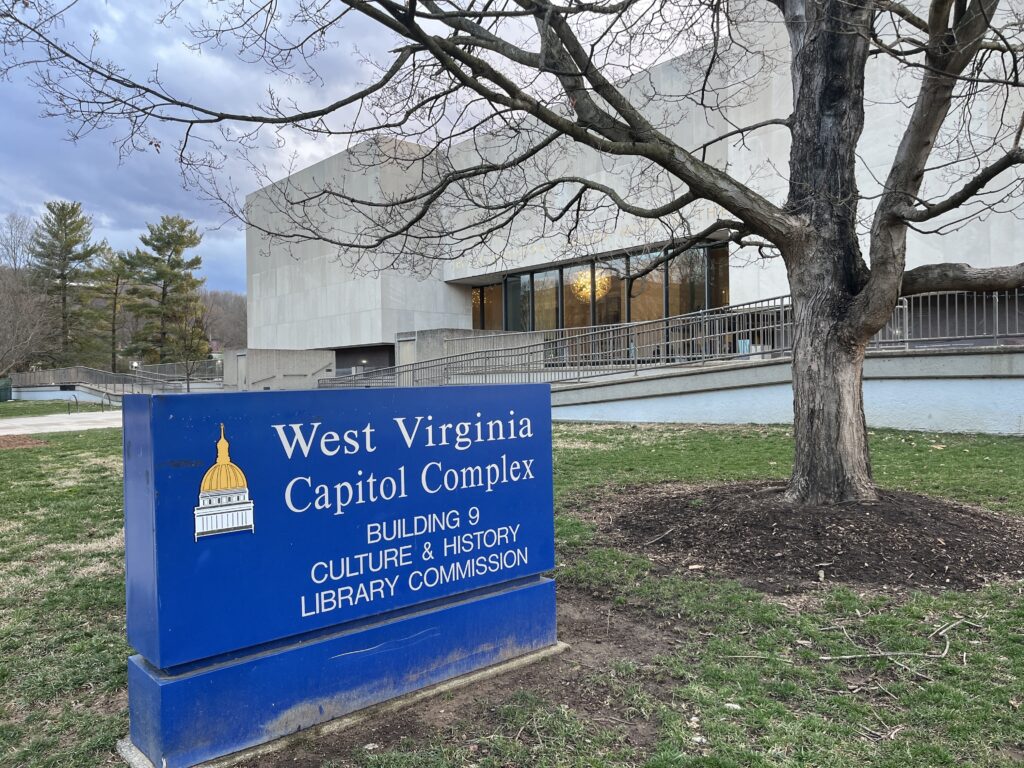 A blue sign with an image of the State Capitol dome reads "West Virginia Capitol Complex Building 9 Culture & History Library Commission." It sits in the grass outside a cement building with tall windows.
