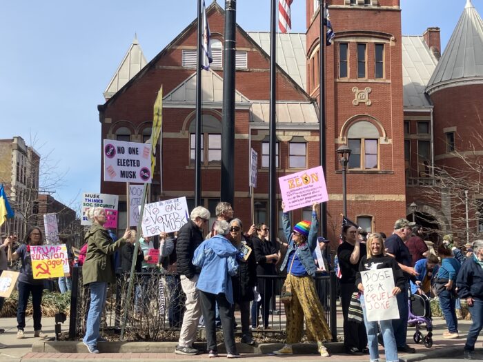 A group holds up homemade signs in front of a red brick building with a prominent bell tower on its right.